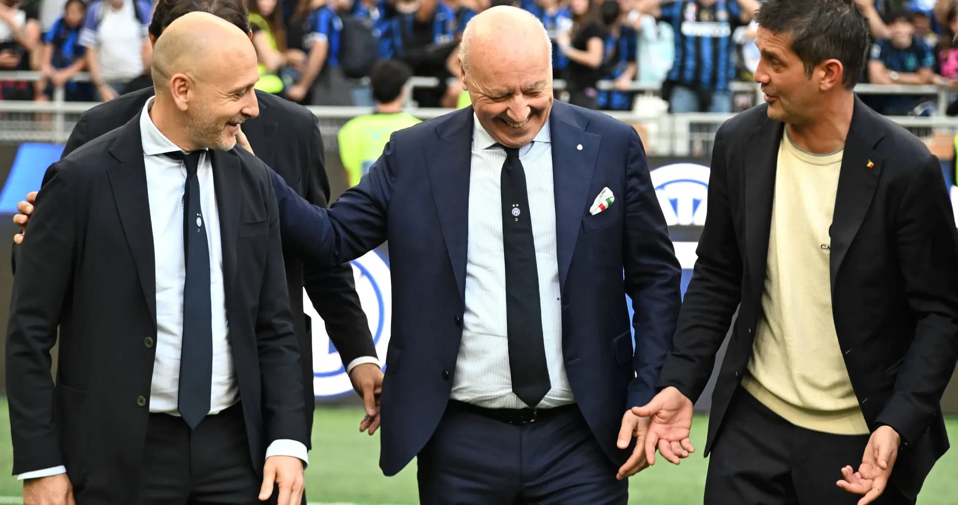 Three men in suits smiling on the pitch Three men in suits smiling on the pitch